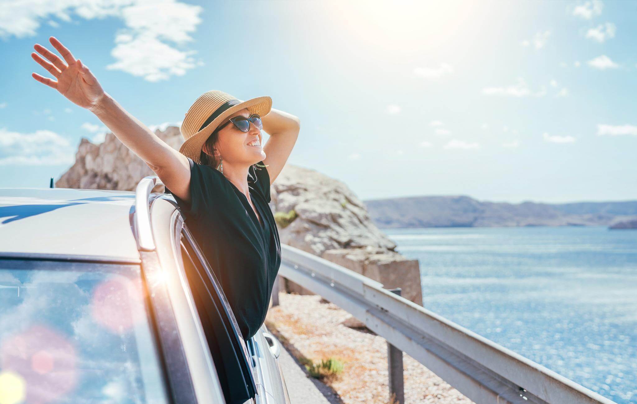 happy traveler woman in hat leaning out of car window with arms wide looking over the water