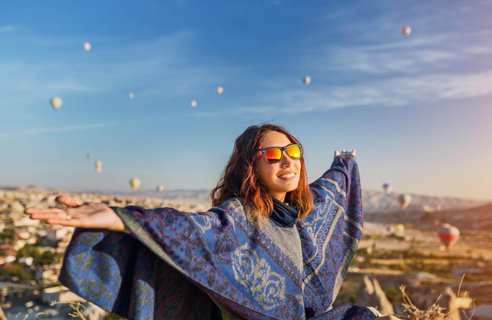 happy female traveler in shawl smiling and extending arms to the sky with hot air balloons in background