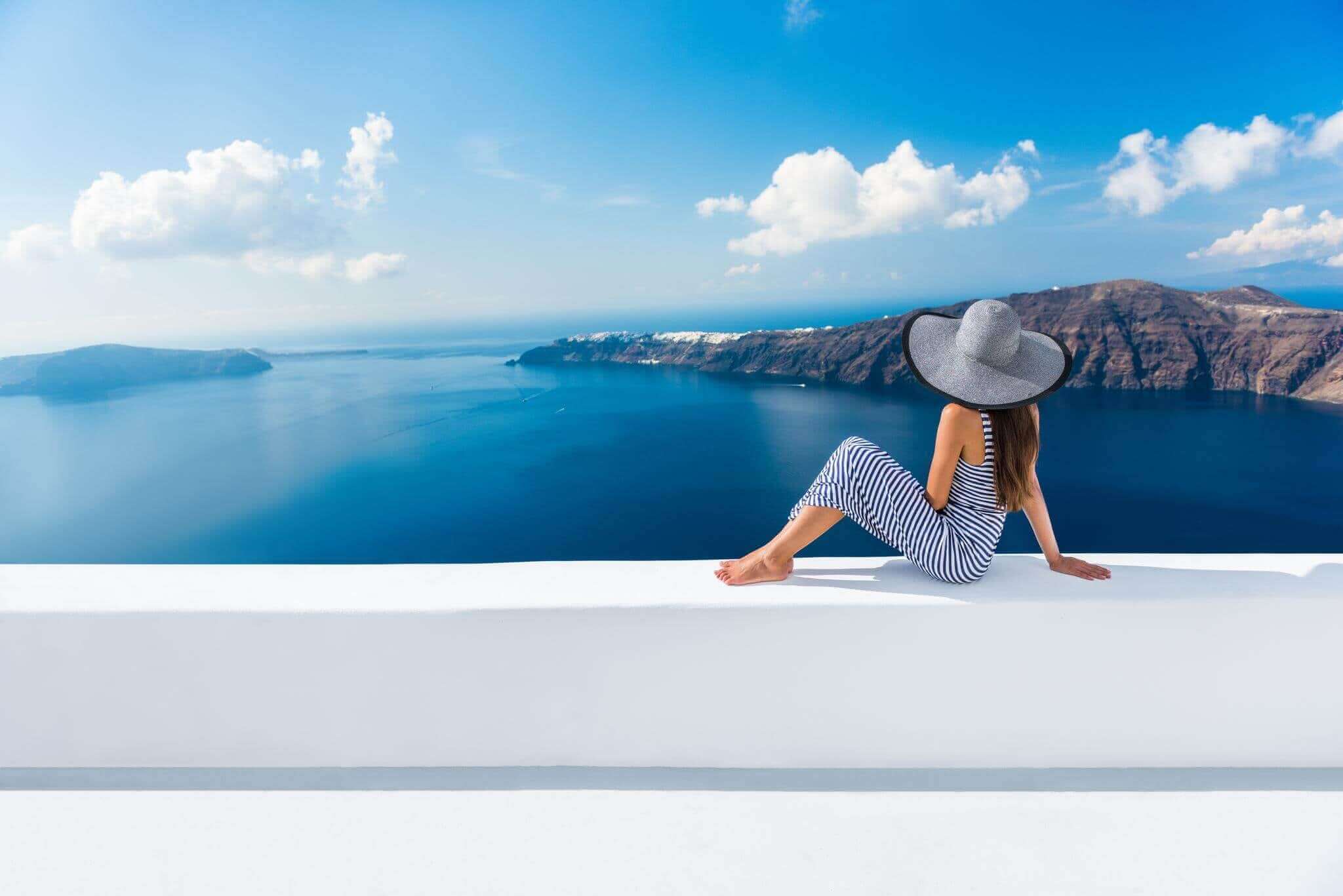 female traveler wearing hat sitting on white ledge overlooking ocean in greek islands