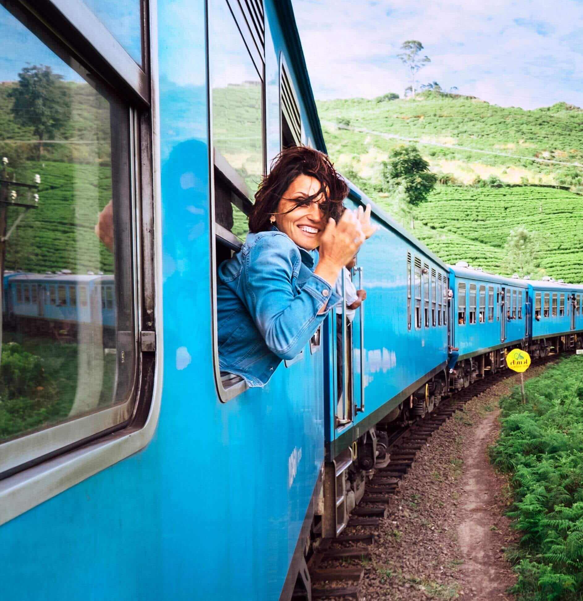 female traveler in jean jacket leaning out window of blue train and smiling as wind blows with green hills in background