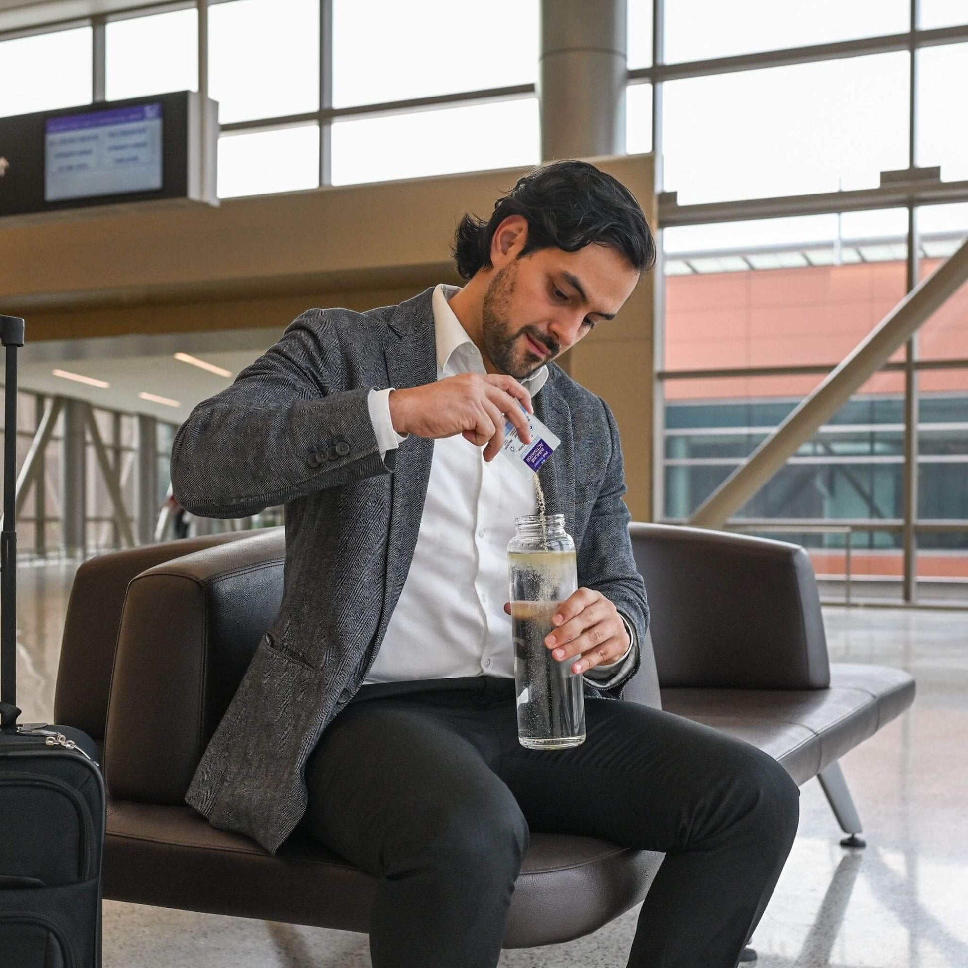 man in suit jacket in airport sitting down and pouring well traveled into water bottle