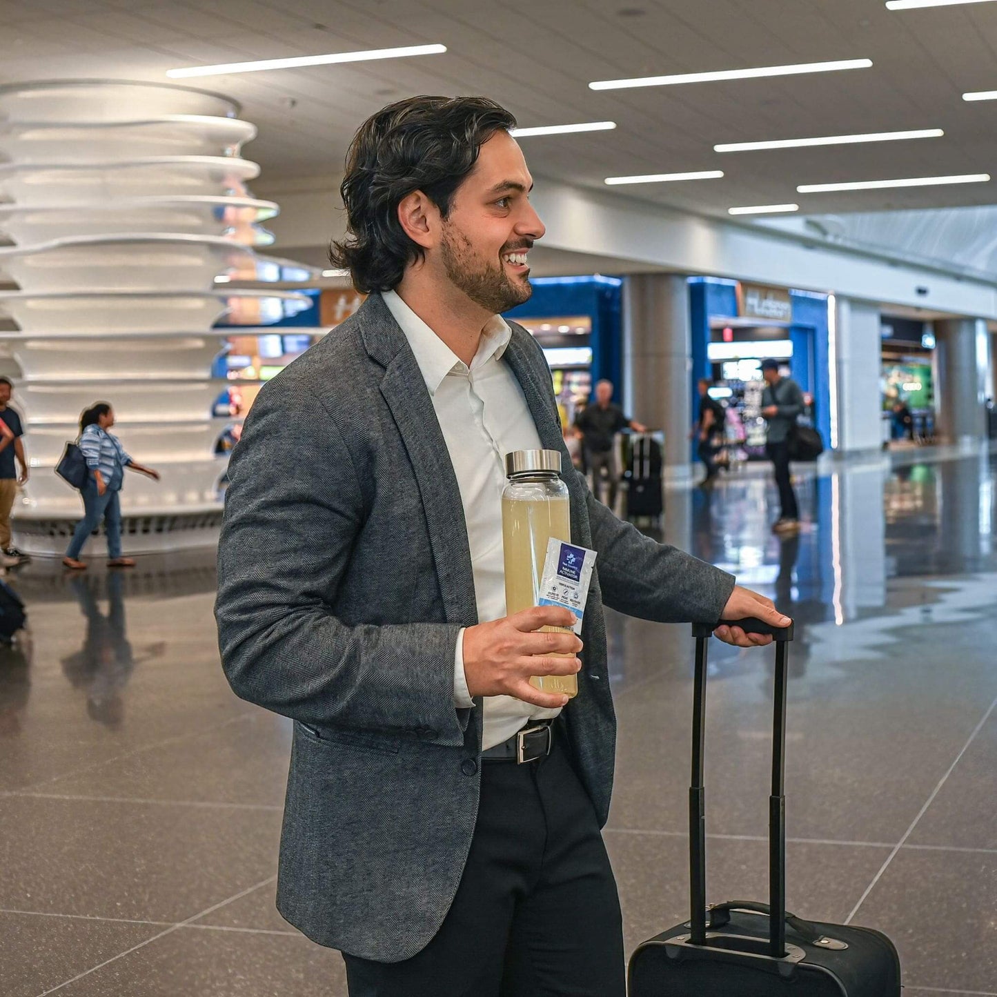 man in suit jacket in airport walking with luggage and holding water bottle with well traveled and packet